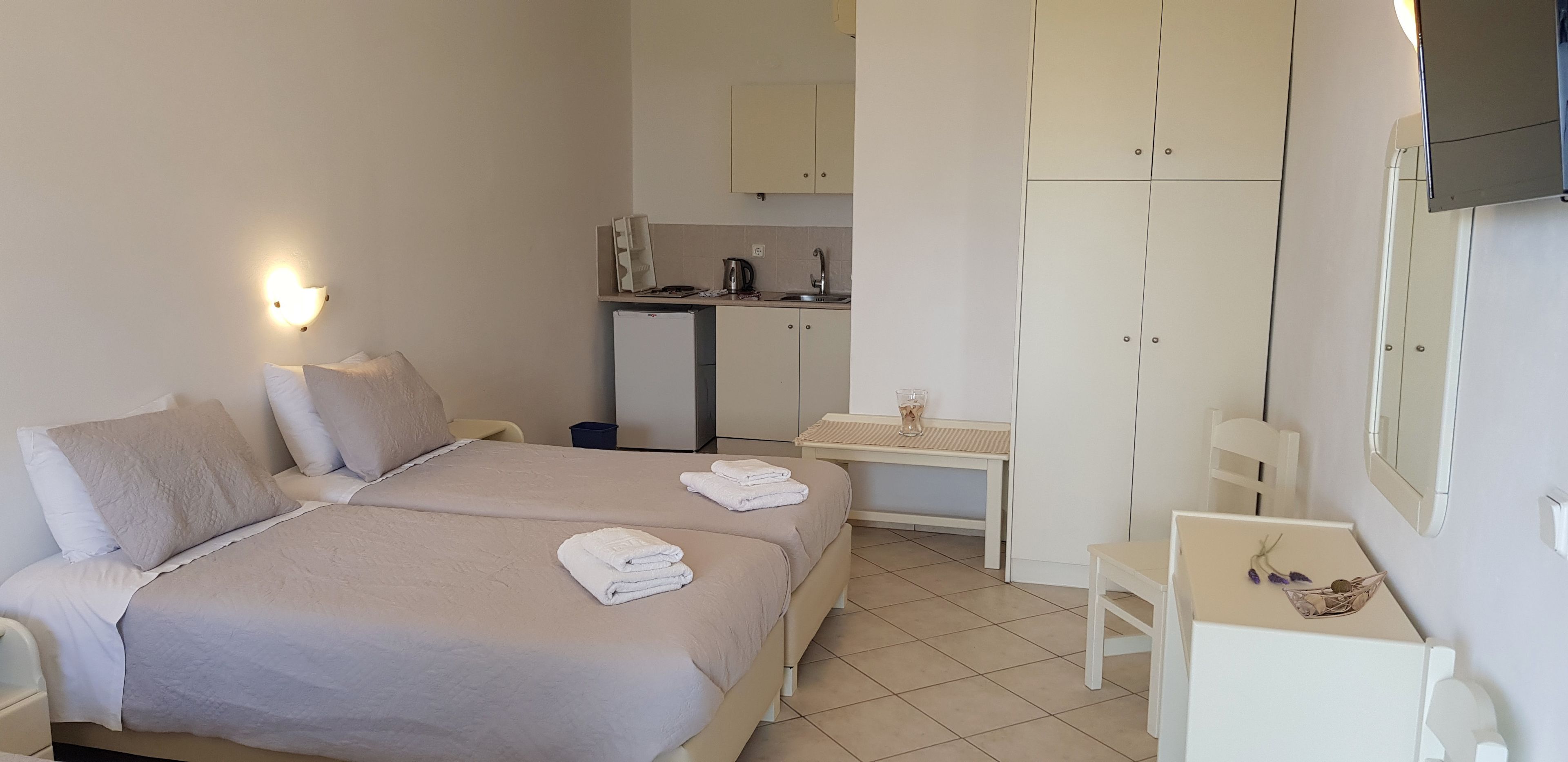 Une chambre d’hôtel avec deux lits, un bureau, une chaise, une petite kitchenette et un téléviseur fixé au mur.