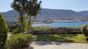 A coastal landscape with a stone wall, a grassy area, and a view of a town and mountains in the background. - Paros Paradise Apartments (Paros)