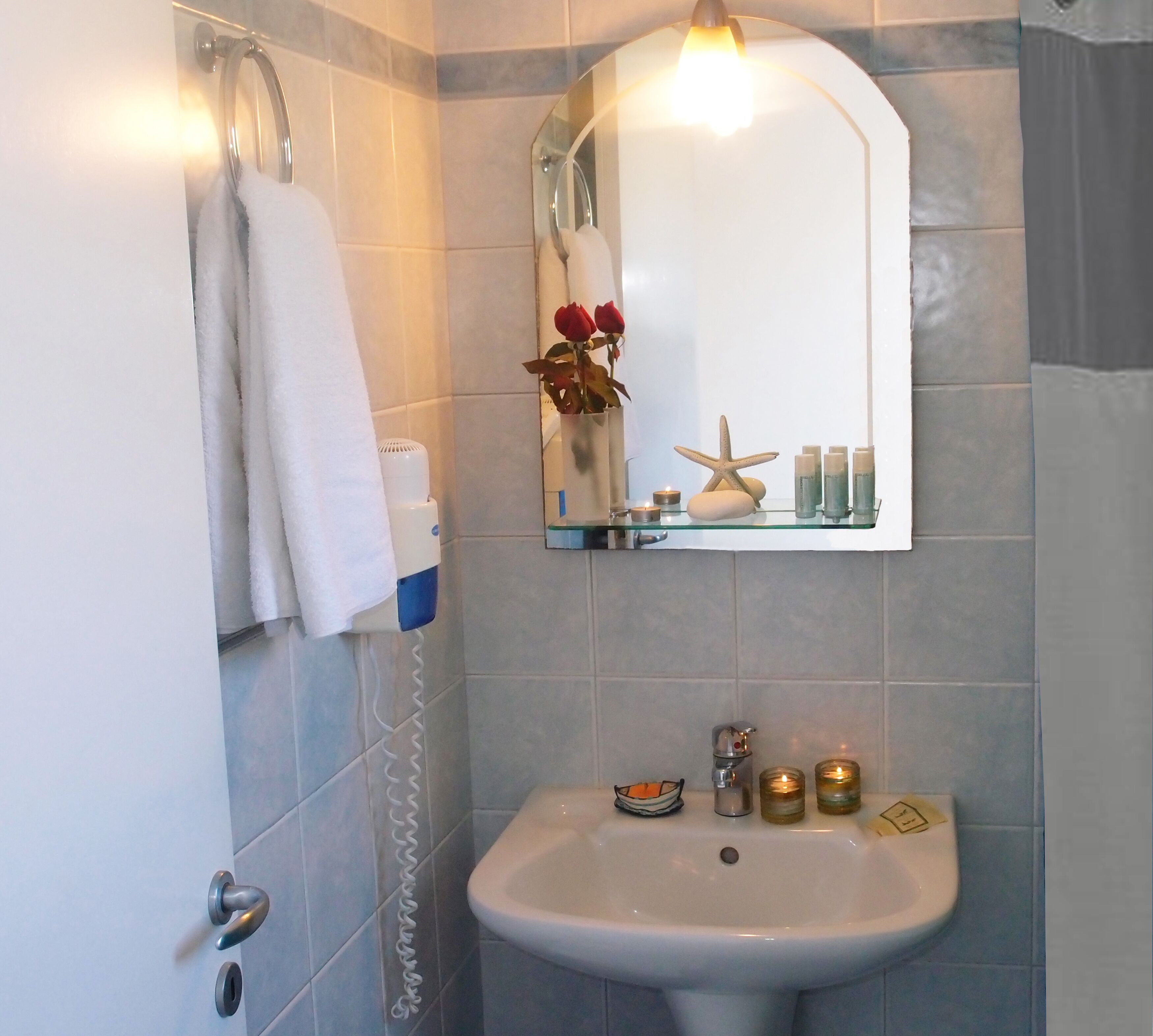 Een badkamer met een witte wastafel, een spiegel, een handdoekrek en een glazen plankje met kaarsen en toiletartikelen.