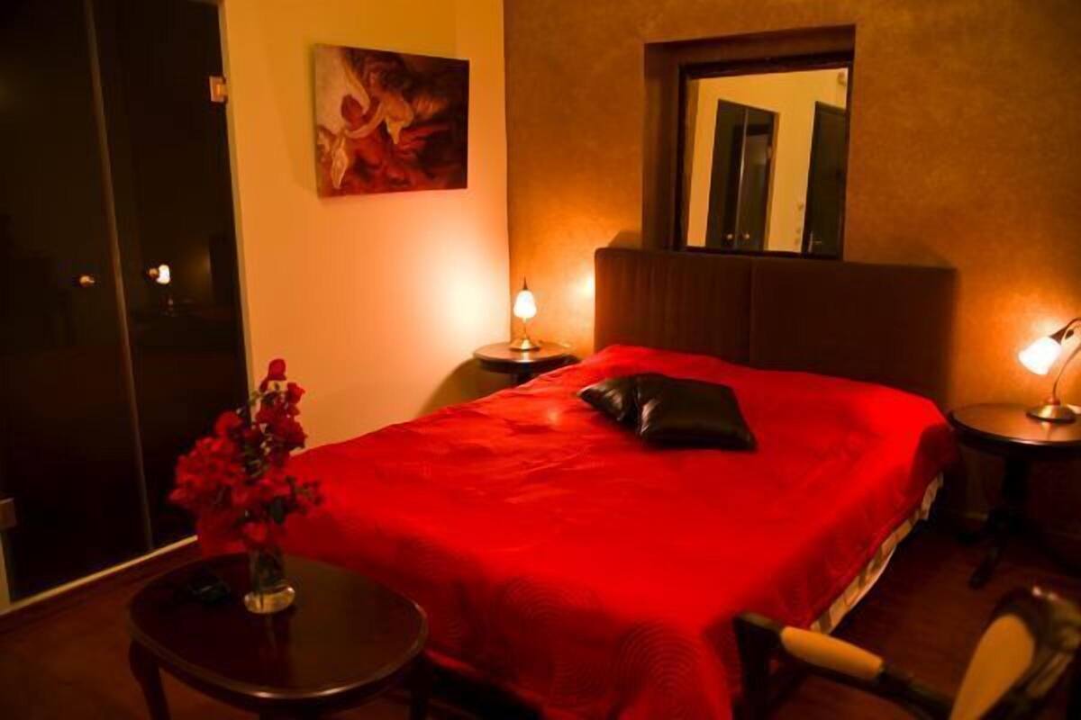 A hotel room with a red bedspread, a painting on the wall, a vase with flowers, and a mirror above the bed.