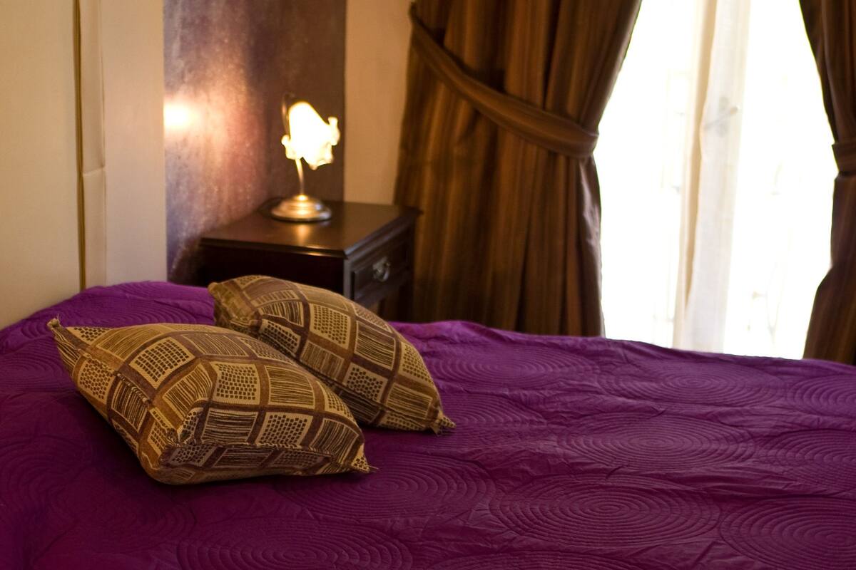 A bed with a purple bedspread and two patterned pillows, a bedside lamp, and a dark nightstand.