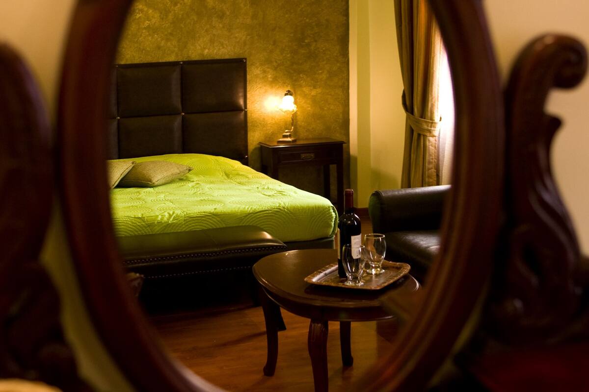 A mirror reflecting a room with a bed, a bedside table with a lamp, and a chair.