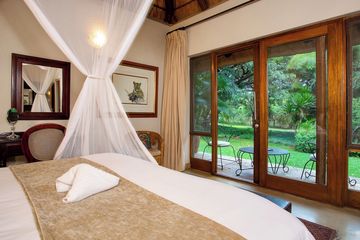 Een slaapkamer met een groot bed, een nachtkastje met spiegel, een stoel en een raam met uitzicht op een tuin.