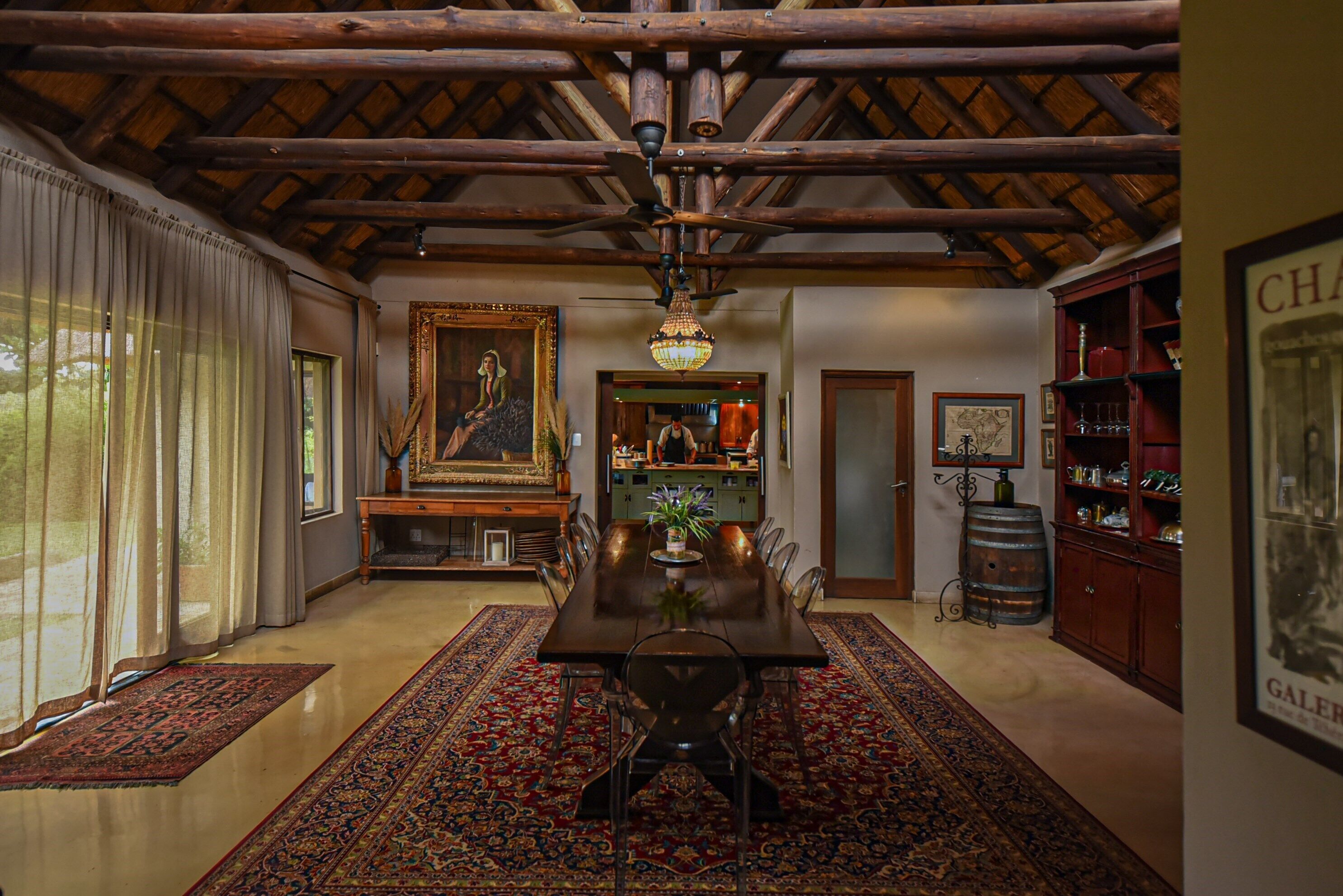 A dining area with a wooden ceiling, a chandelier, a large painting, and a bar area with shelves and a barrel.