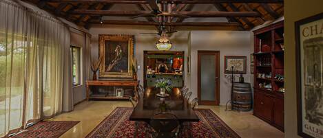 A dining area with a wooden ceiling, a chandelier, a large painting, and a bar area with shelves and a barrel.