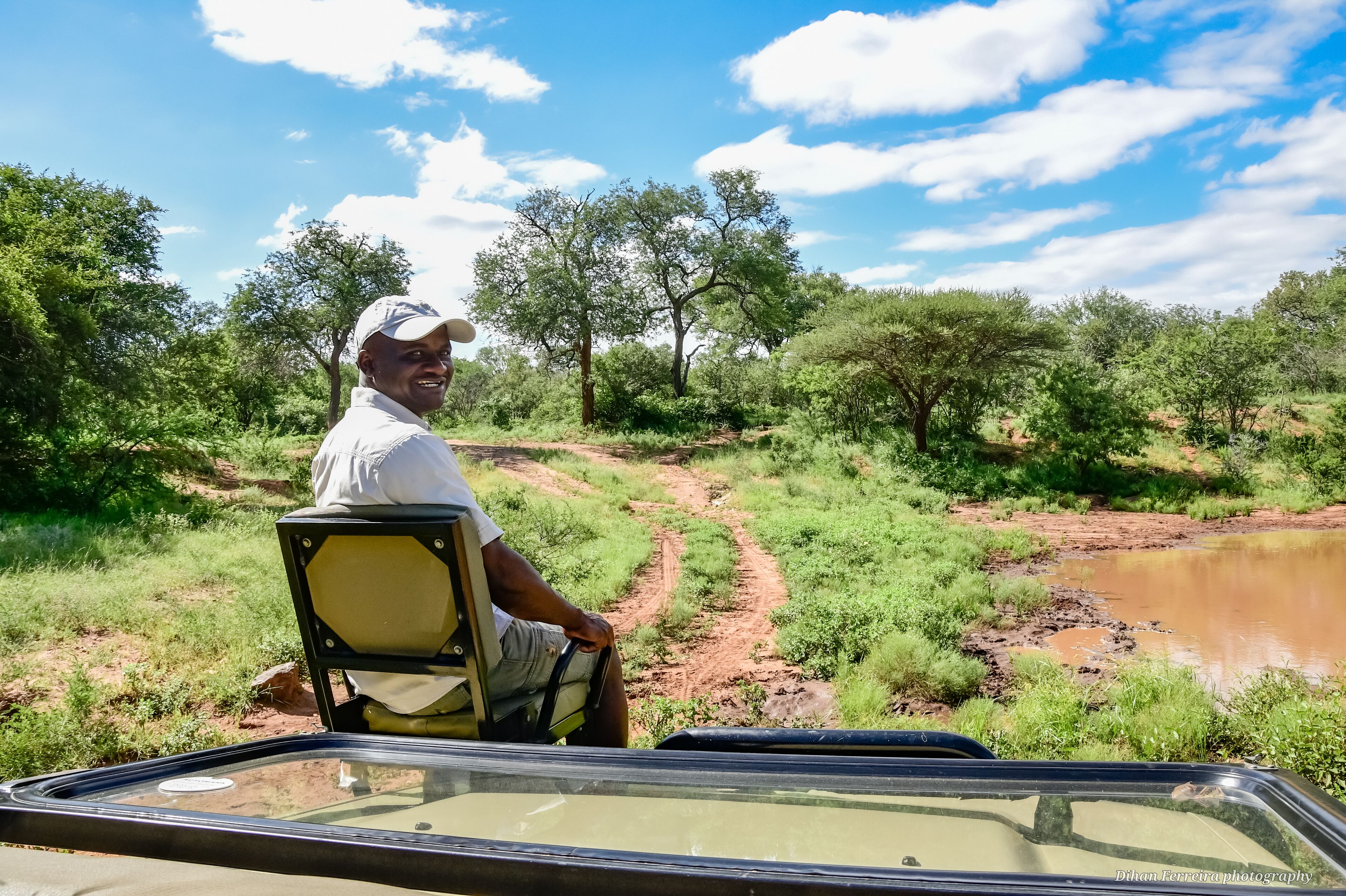 A person seated in a vehicle with a view of a dirt path and a water body surrounded by greenery.