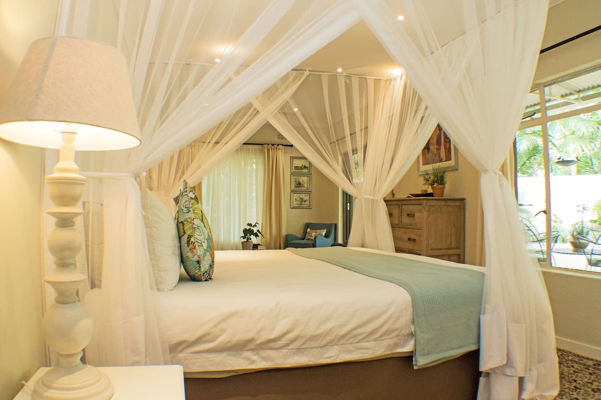 Ein Schlafzimmer mit einem Bett, einer Nachttischlampe, einem Sessel, einem Schminktisch und einem Fenster mit Vorhängen.
