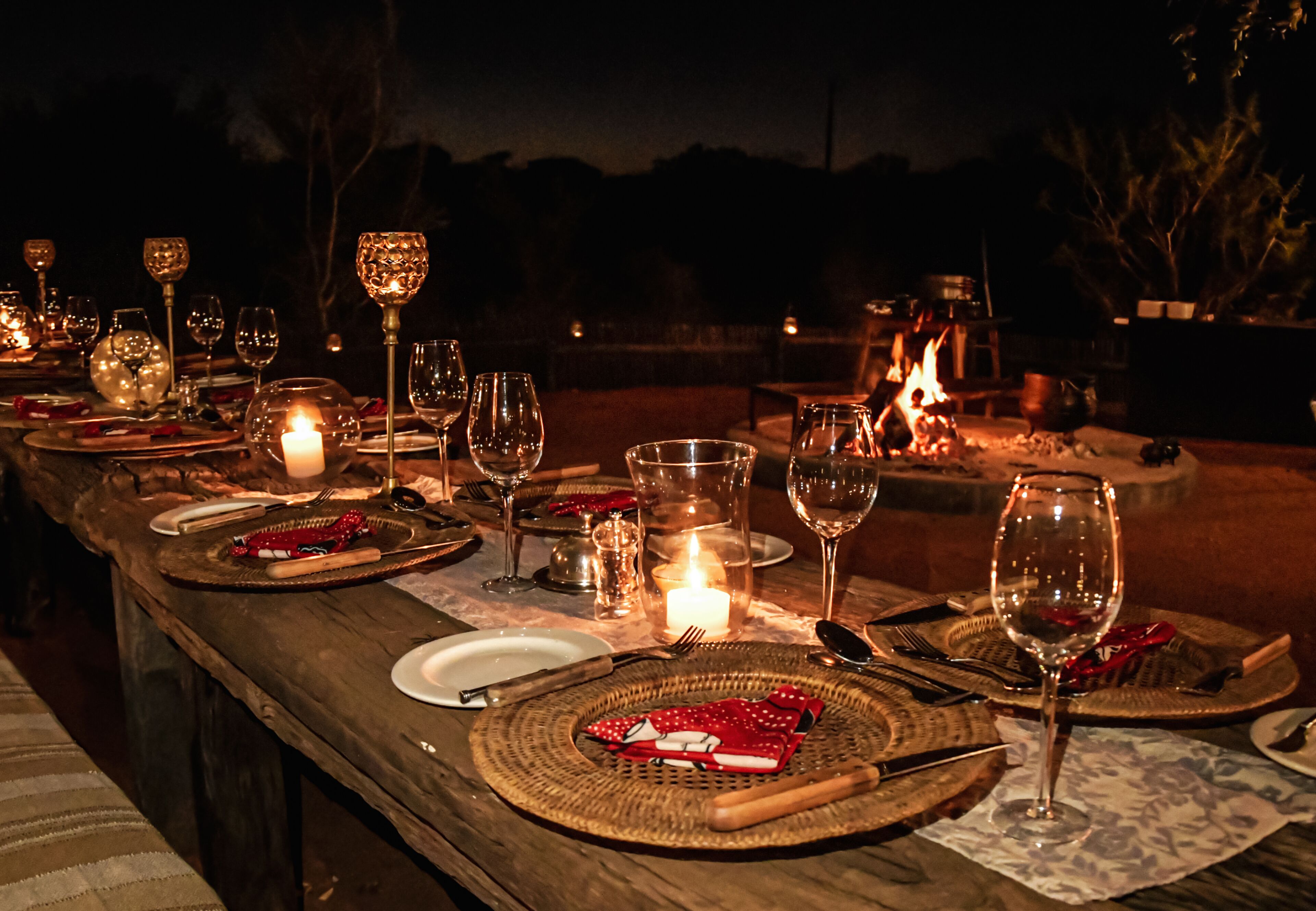 An outdoor dining table set with various glassware, plates, and cutlery, surrounded by a fire pit and candles, indicating a cozy evening meal setting.