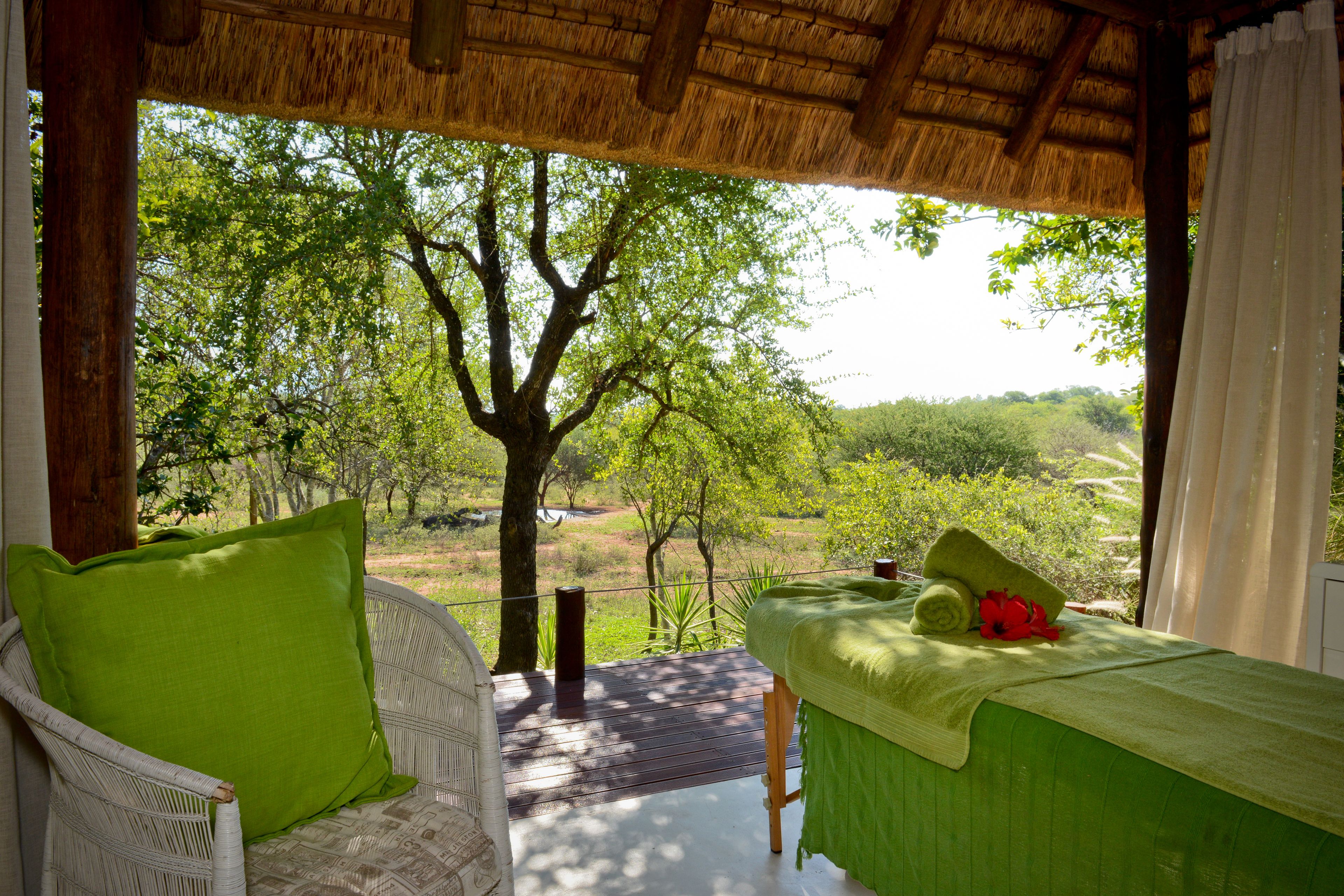 A room with a wooden ceiling, a green bed, a white chair, and a view of trees and greenery outside.