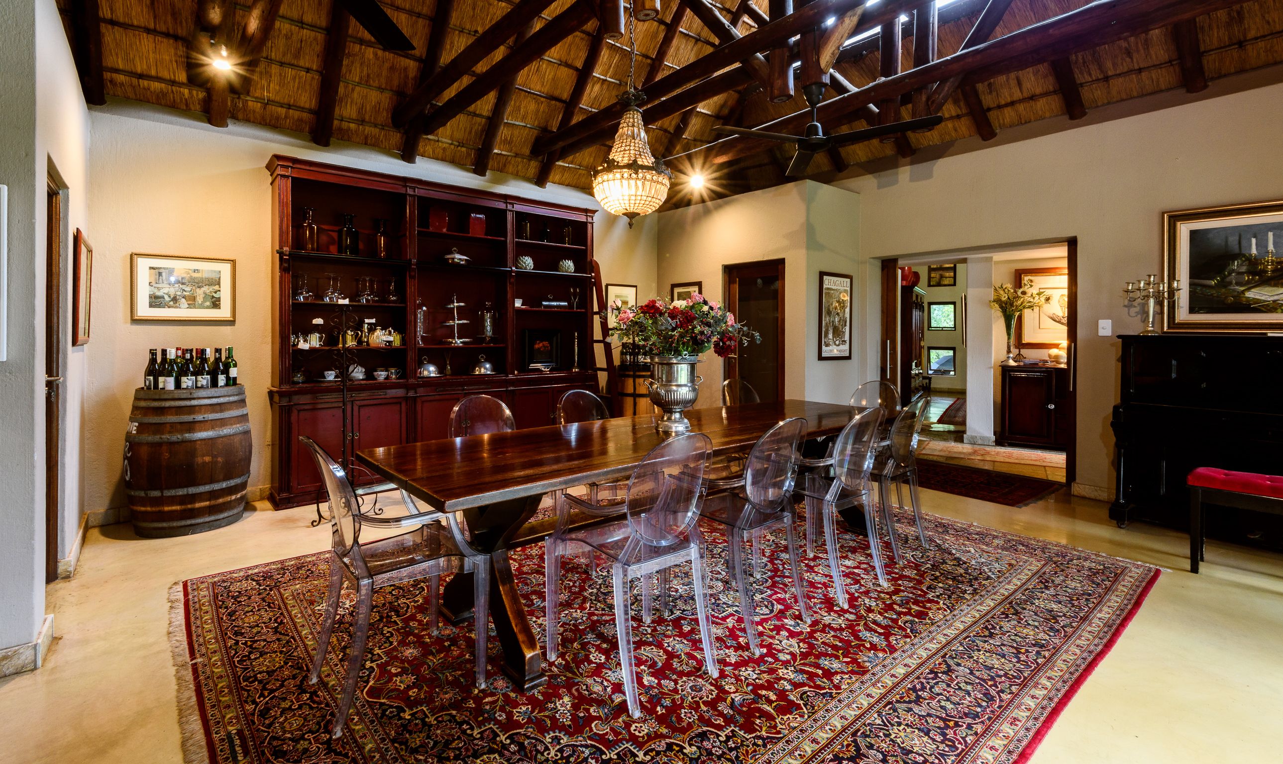 A dining area with a wooden table, clear chairs, a red patterned rug, a wooden cabinet with glassware, and a hanging chandelier.