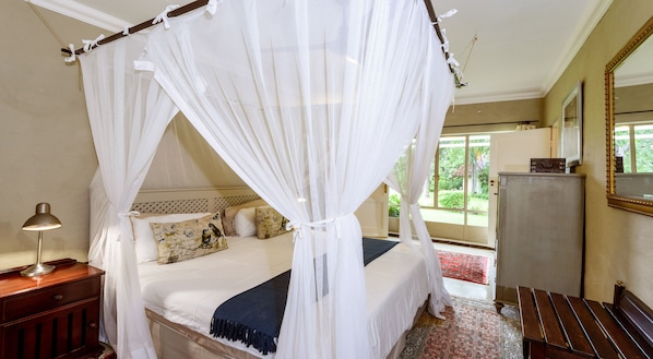 Un dormitorio con cama con dosel, una lámpara de noche, una mesita de noche, un aparador, un espejo y vistas al verdor exterior.