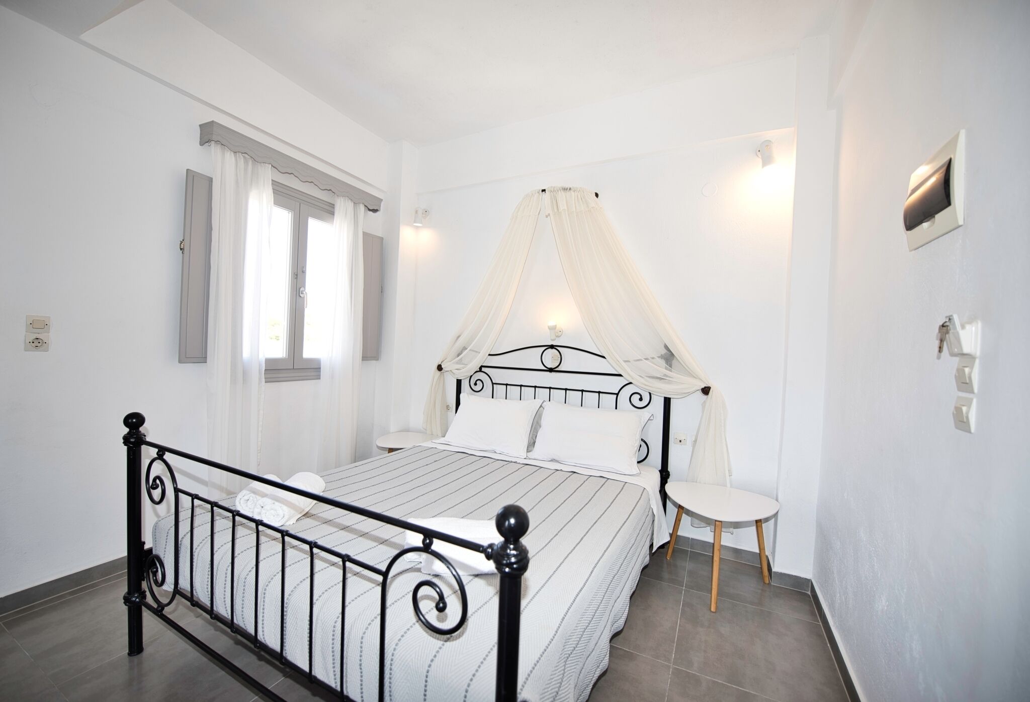 Un dormitorio con una cama, una mesita de noche, una silla, una ventana con cortinas y una lámpara fijada a la pared.