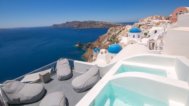 A seaside hotel balcony with white walls, blue domed roofs, and a view of the sea and distant hills.