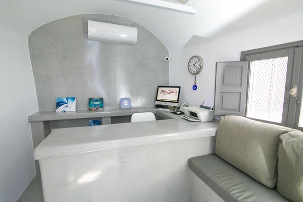 A reception area with a counter, a computer, a clock, and a couch. - Theodora Suites - Adults Only (Santorini)
