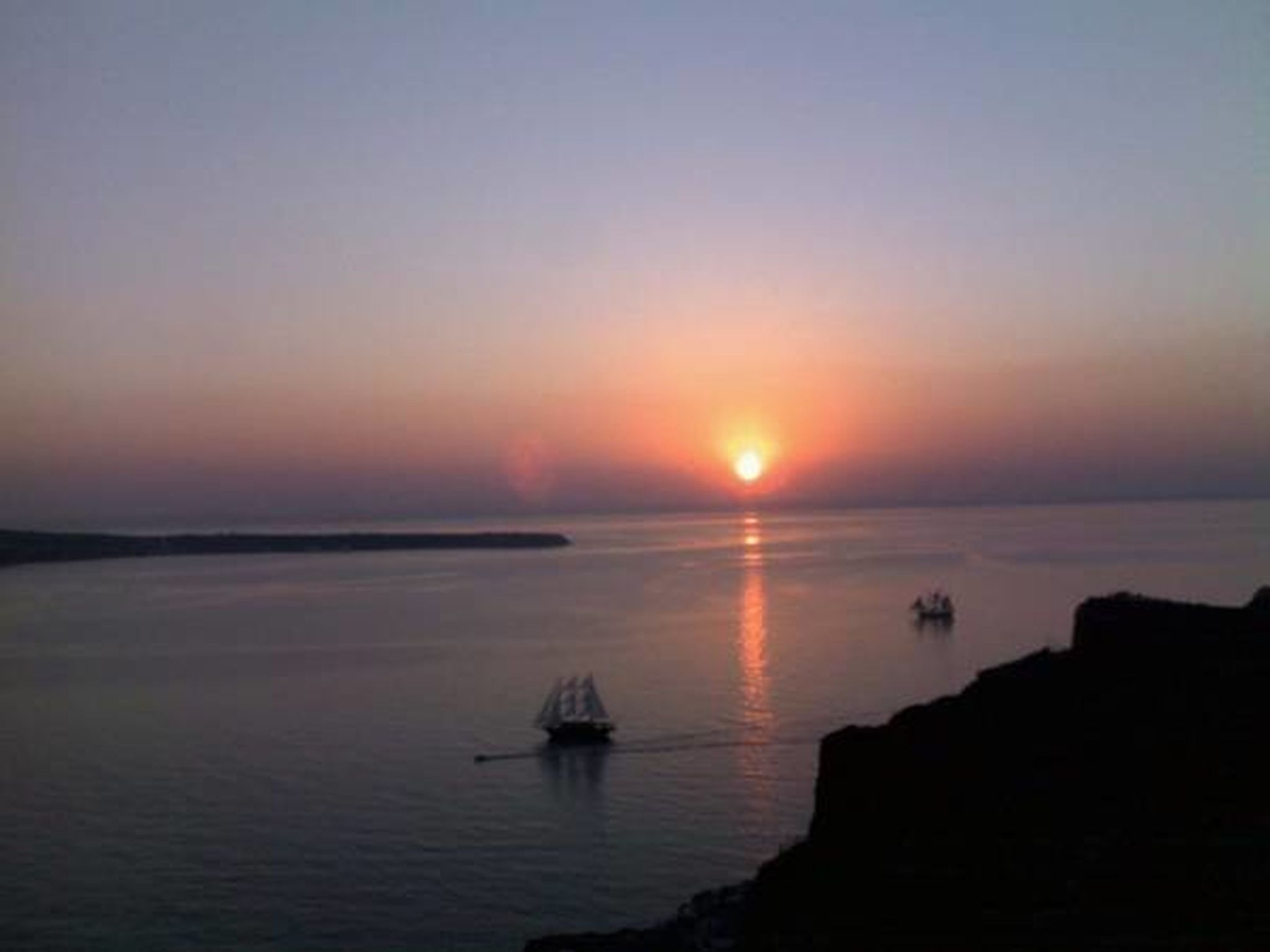 A sunset over a calm sea with a sailboat and a distant shoreline.