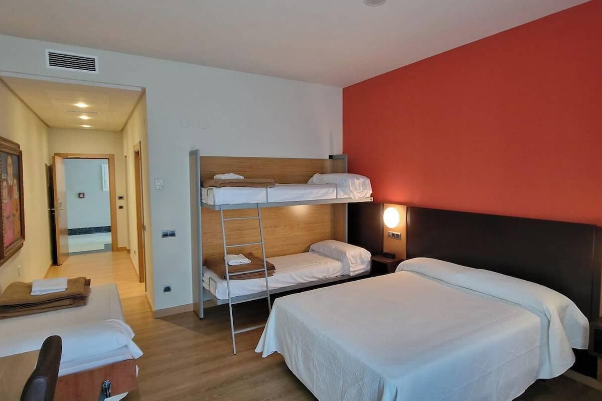 A hotel room with bunk beds, a desk, a chair, a bedside table, and a wall-mounted lamp.