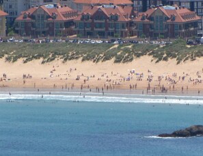 A beach with people and cars, and a row of buildings with red roofs in the background.