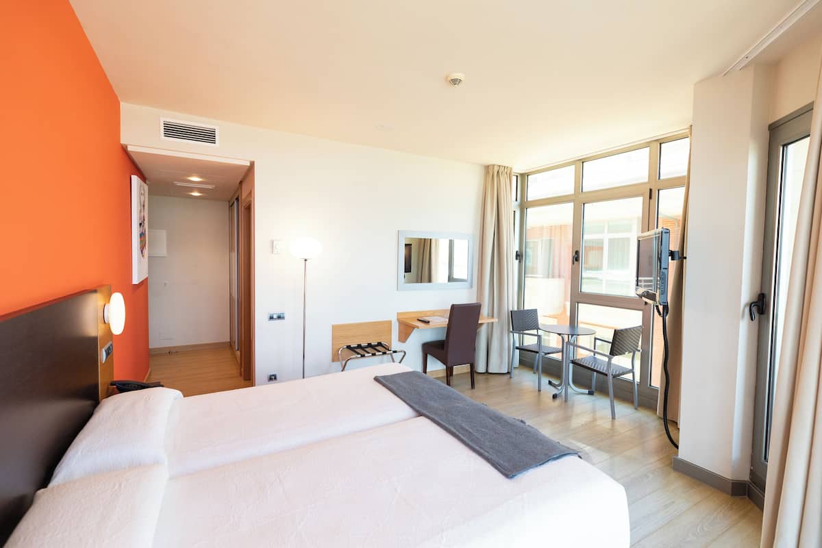 A hotel room with a large bed, a desk, a chair, and a view of the outside through large windows.