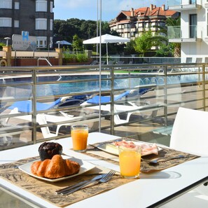 A dining table with croissants, juice, and a plate of food, overlooking a pool with lounge chairs and umbrellas.