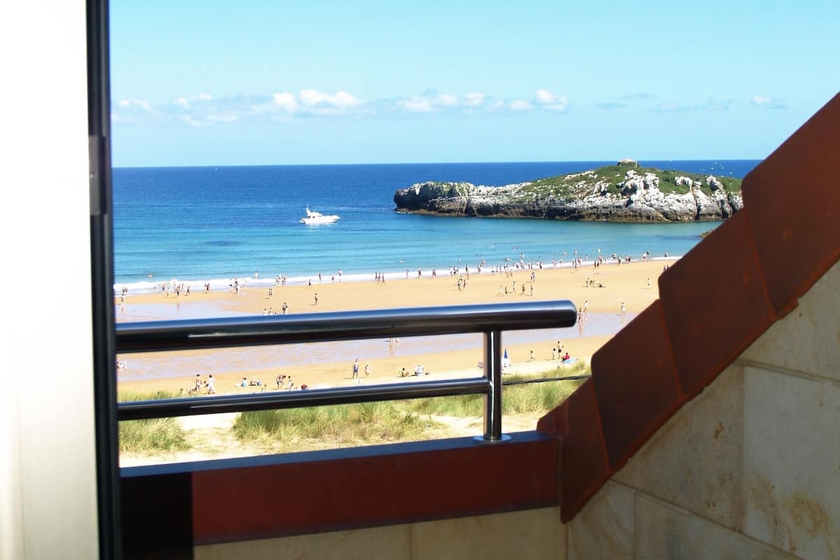 A view from a window overlooking a beach with a rocky outcrop and a boat on the water.