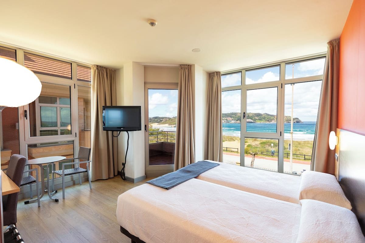 A hotel room with two beds, a TV, a balcony with a view of the beach, and a dining area.