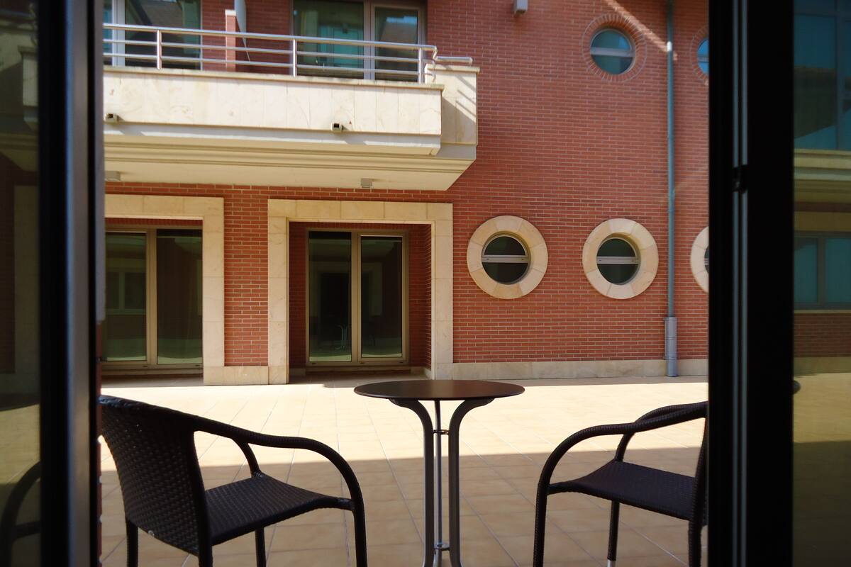 A patio area with two chairs and a table, a red brick building with windows and a balcony in the background.