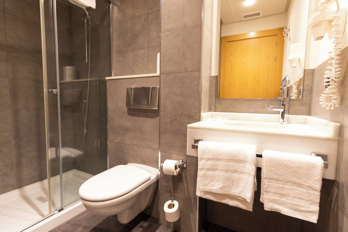 A modern bathroom with a glass-enclosed shower, a toilet, a sink with two towels, and a mirror with a wooden door in the background.