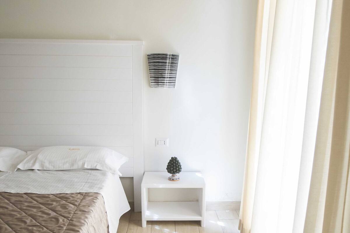 A bedroom with a bed, a nightstand, a wall-mounted shelf, and a window with curtains.