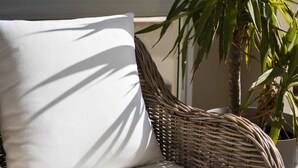 A wicker chair with a white cushion, a potted plant, and a view of buildings through a window. - Hotel Sole (Lampedusa)