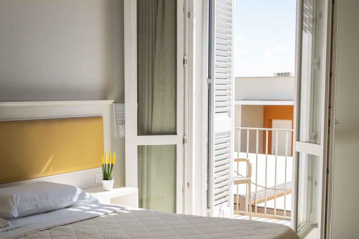 A bedroom with a bed, a nightstand, a window with curtains, and a view of an outdoor balcony.