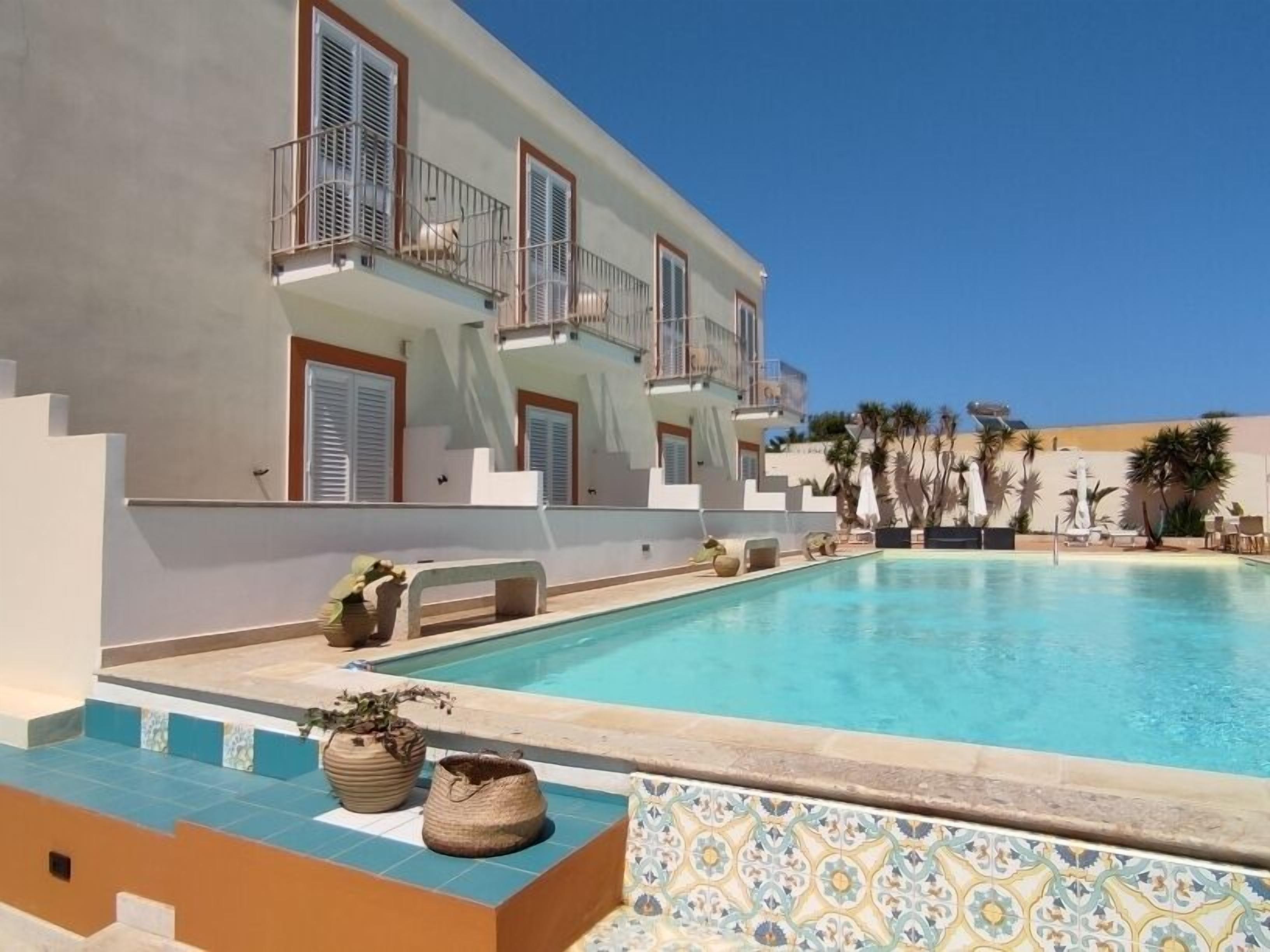 A pool area with a clear blue pool, a white building with balconies, and a tiled border.