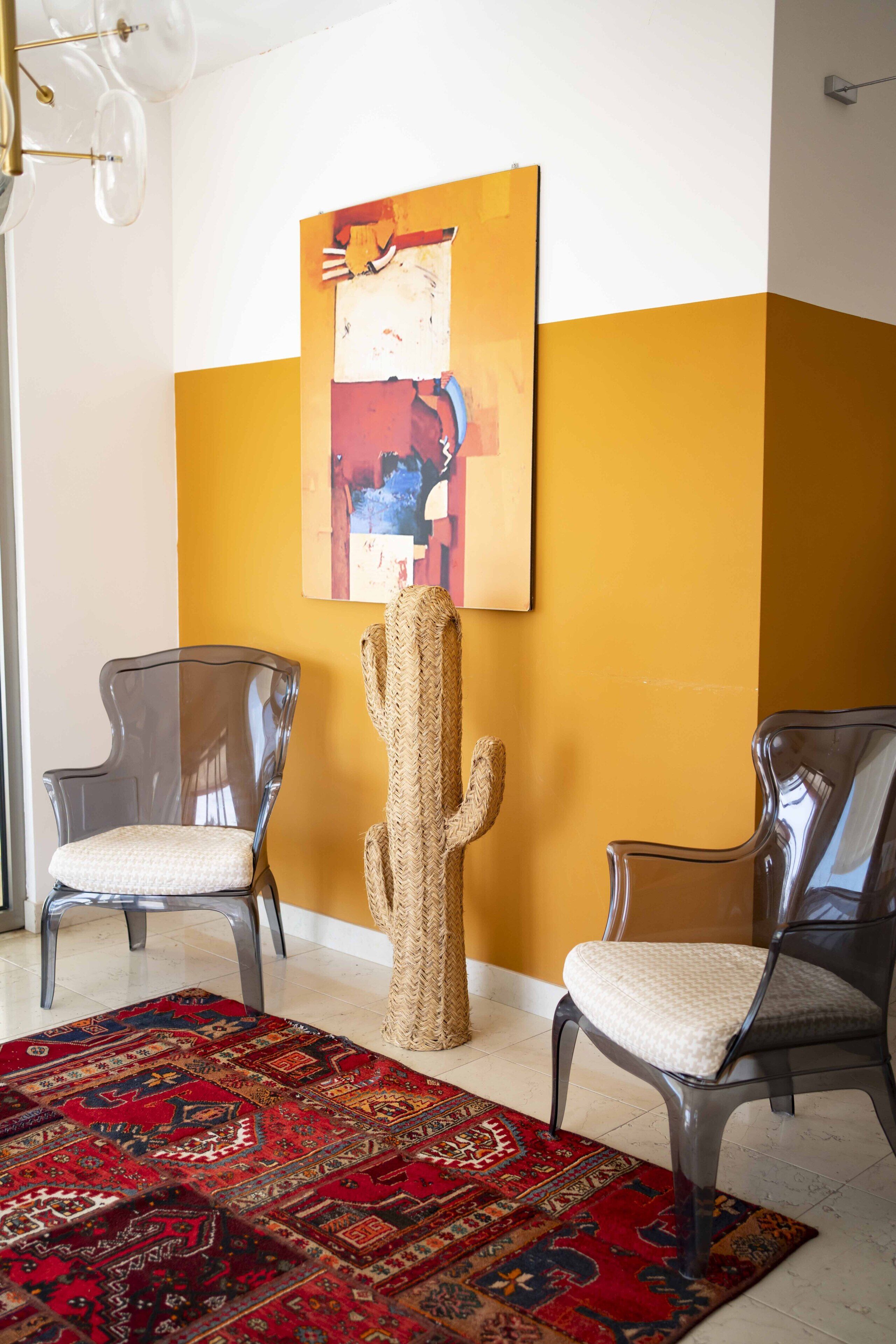Quarto com duas cadeiras, uma cadeira de vime, um tapete vermelho estampado e um grande quadro abstrato na parede.