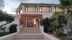 A modern building with a staircase leading to a door, surrounded by various plants and a palm tree. - Hotel Sole (Lampedusa)