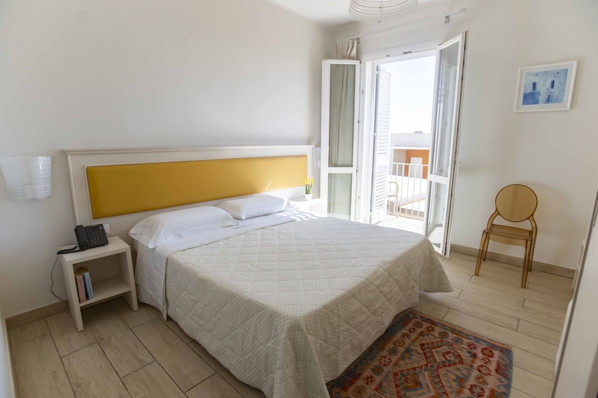 A hotel room with a bed, bedside table, a chair, a small table with a vase, and a view of the outside.