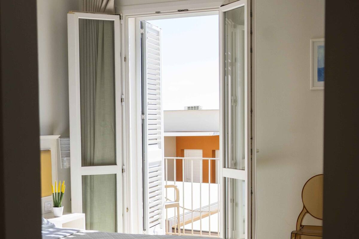 A bedroom with a bed, a chair, a desk, a window, and an open door leading to a balcony.