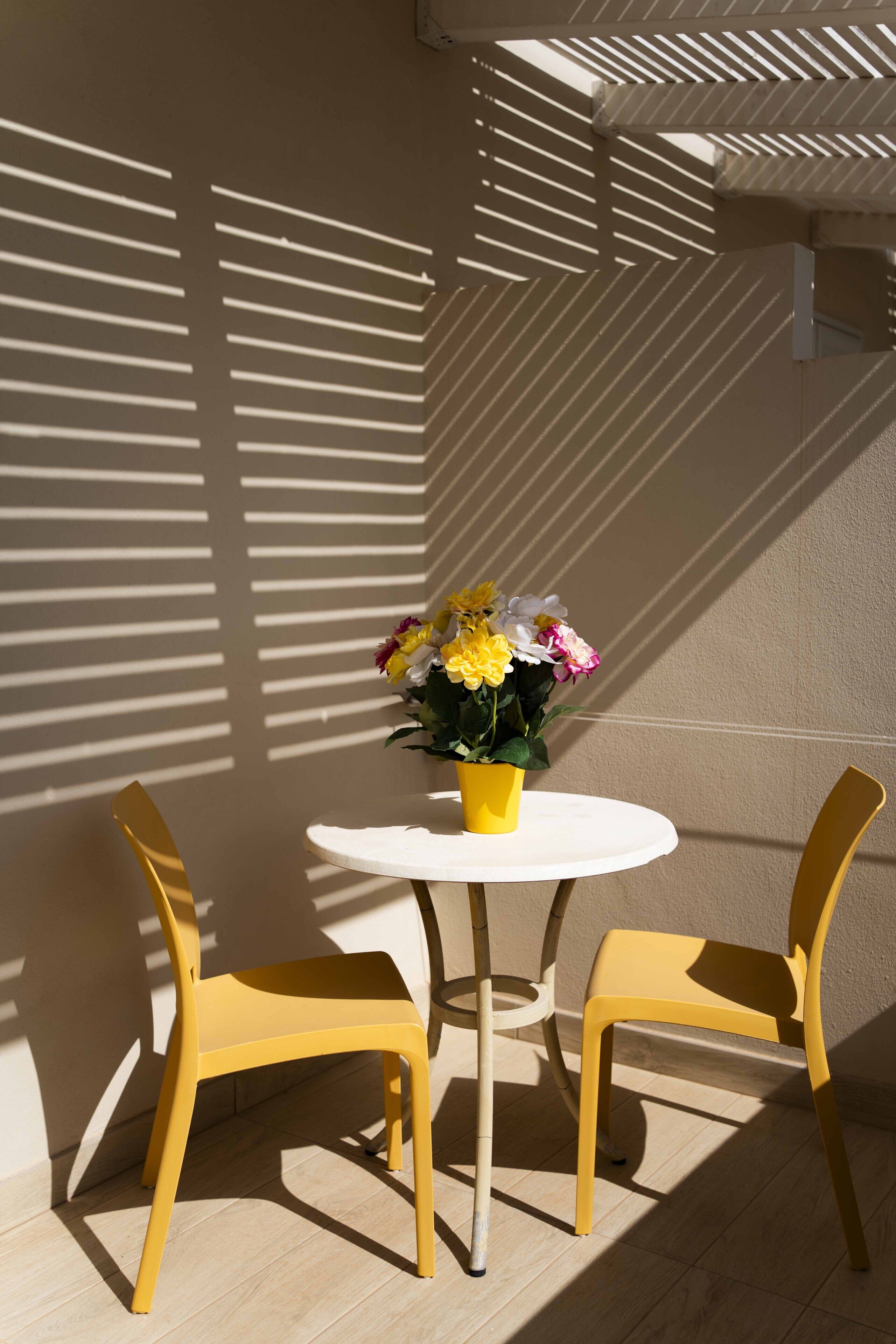 Ein runder Tisch mit einer gelben Blumenvase, zwei gelben Stühlen und einer weißen Wand mit Schatten.
