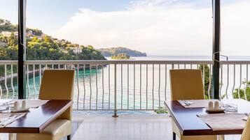 A dining area with a view of a coastal landscape, including a body of water and a hillside with buildings.