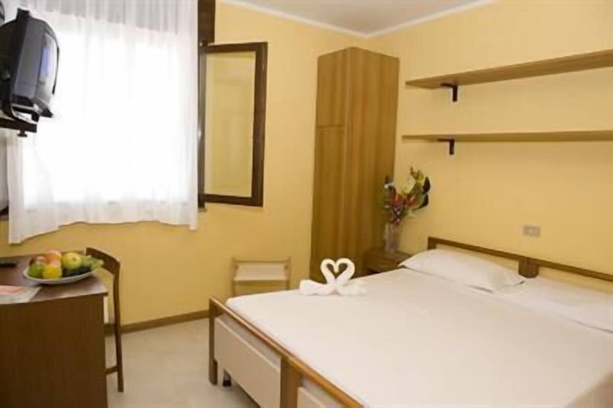 A hotel room with a bed, a desk with a fruit bowl, a mirror, a vase with flowers, and a television.