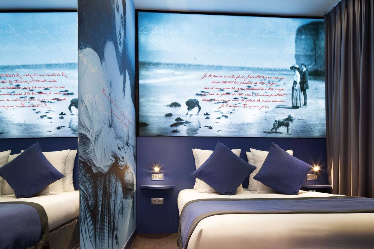 A modern hotel room with a large wall mural of a beach scene, a neatly made bed with blue and white bedding, and a small bedside table with a lamp.