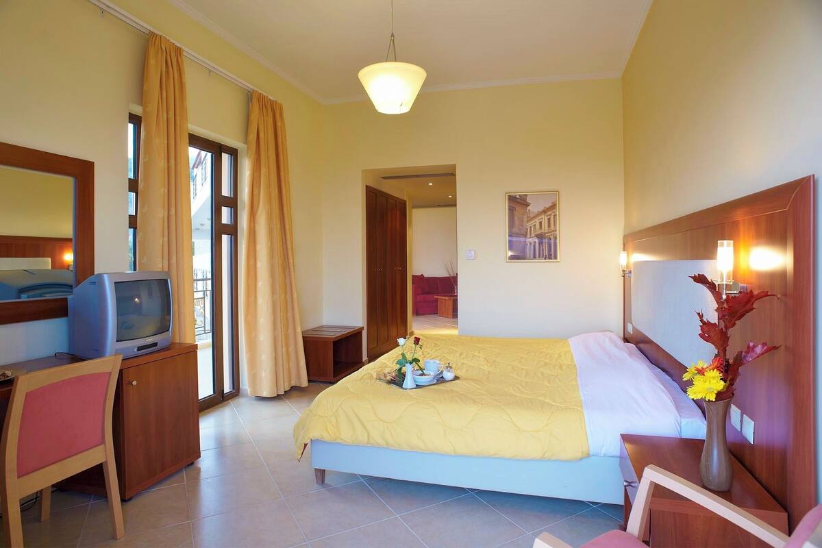 Quarto de hotel com uma cama, uma escrivaninha com televisão, um vaso com flores e vista para o exterior.