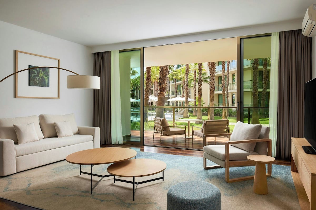 A modern hotel room with a sofa, two round tables, a TV, and a large window with a view of palm trees and a pool.