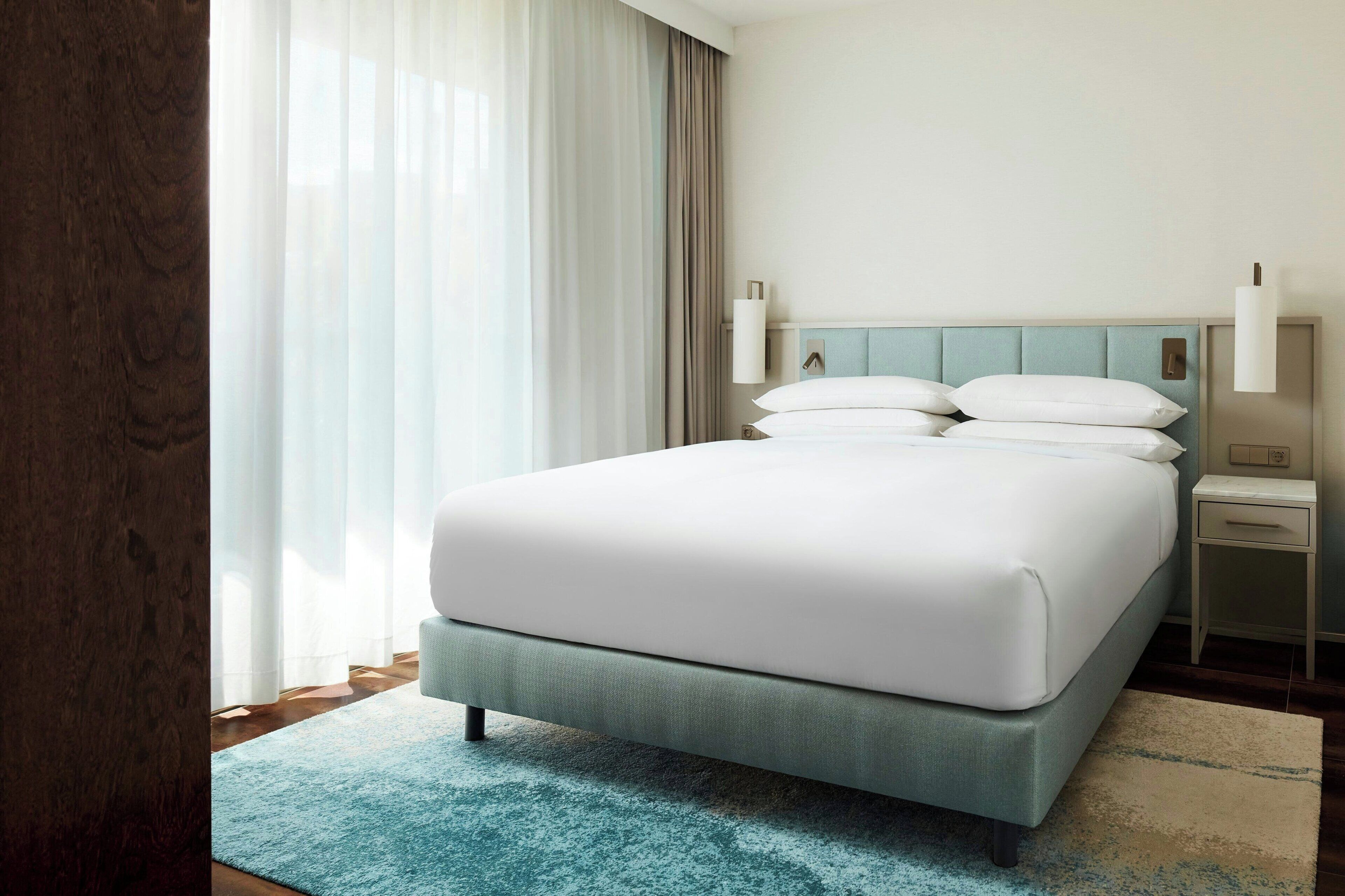Quarto de hotel com uma cama grande, mesa de cabeceira, abajur e vista para o exterior através de cortinas translúcidas.