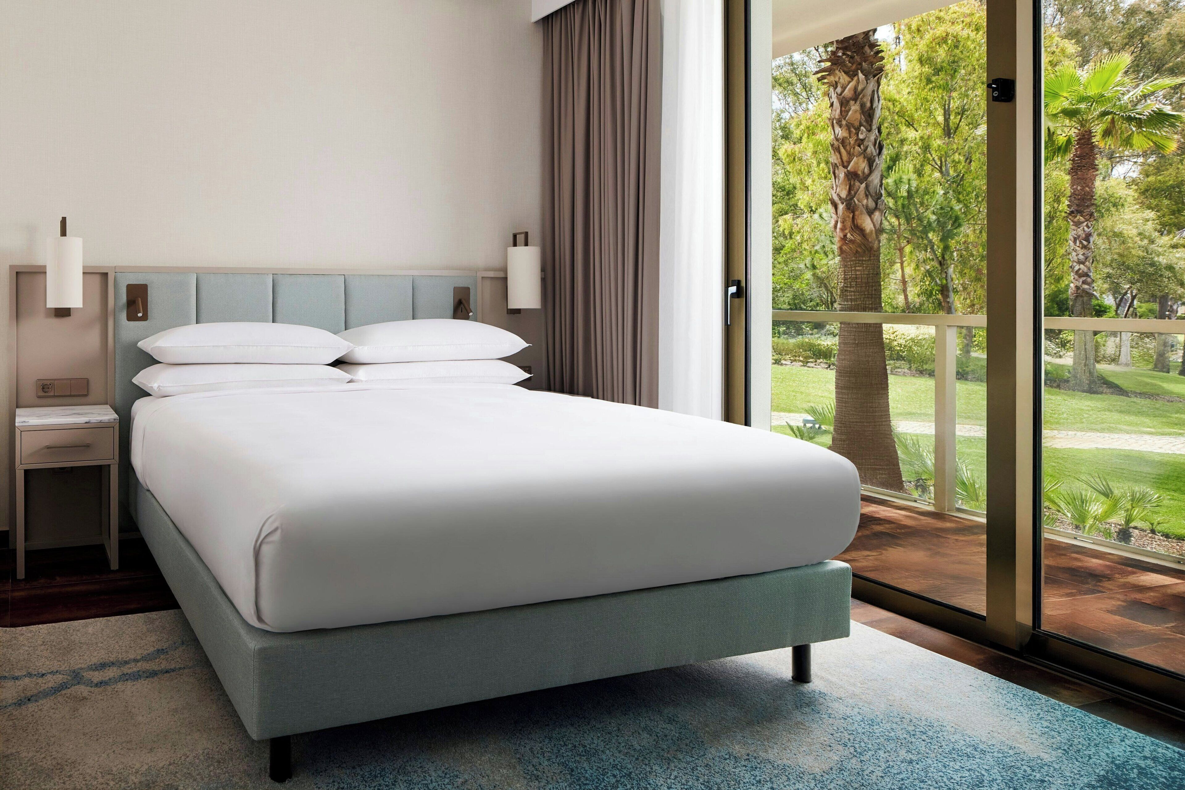 A modern hotel room with a large bed, a nightstand, and a view of a lush garden through a sliding glass door.