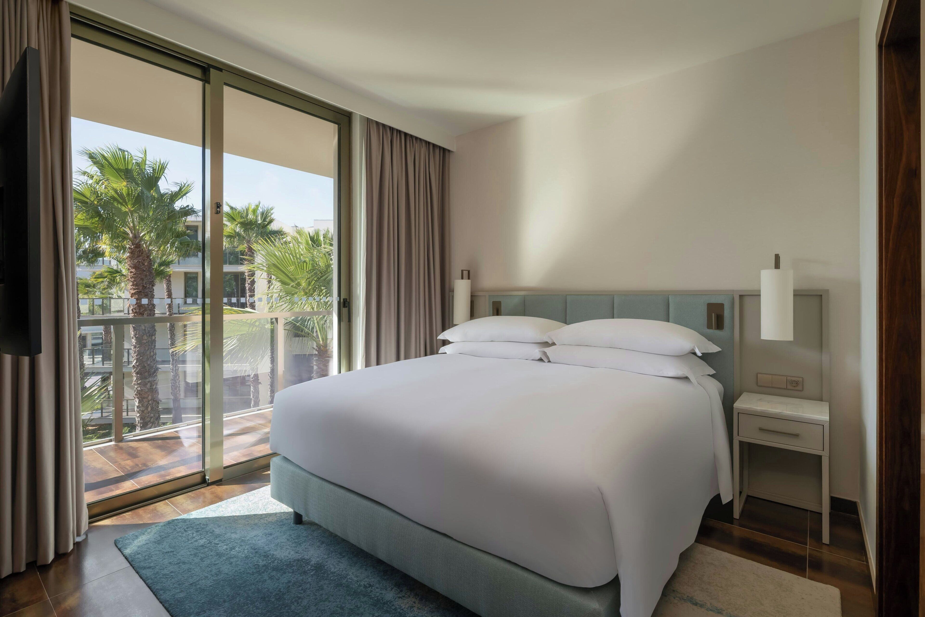 A hotel room with a large bed, a bedside table, a TV, and a view of palm trees and buildings through the window.