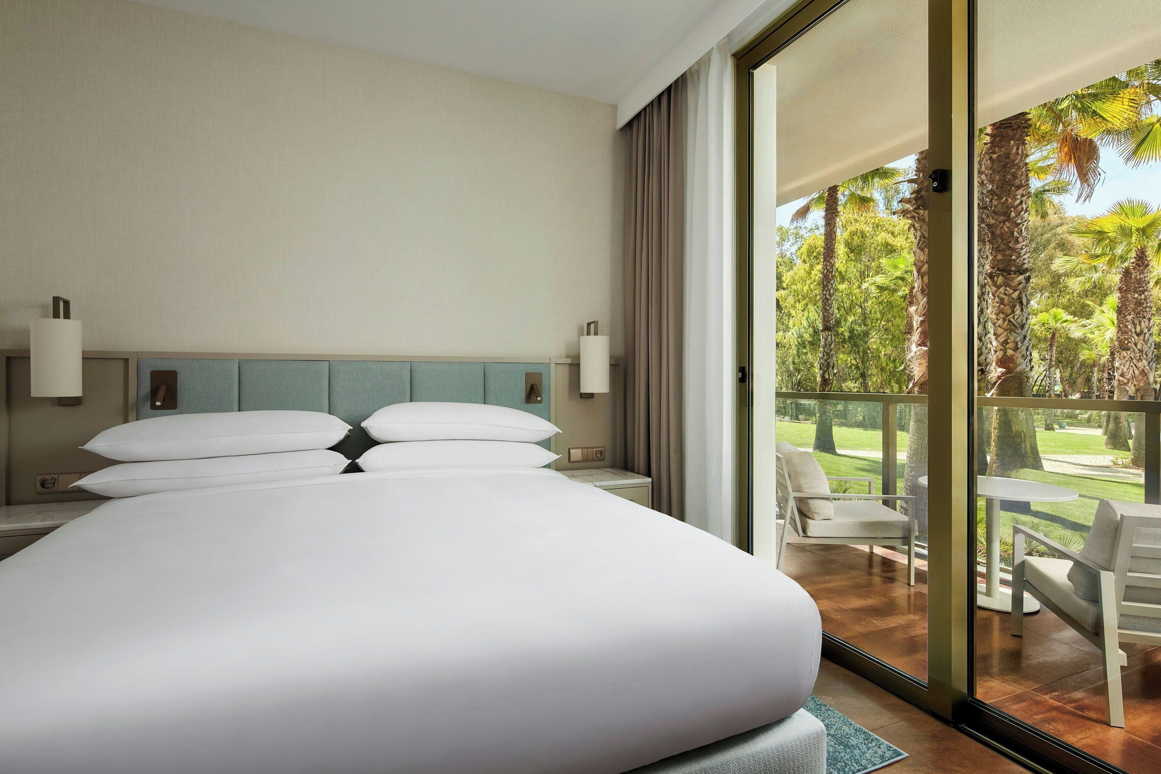 A hotel room with a large bed, a nightstand, a lamp, and a view of a balcony with outdoor furniture.
