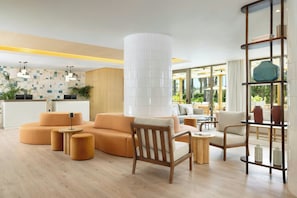 A modern hotel lobby with a wooden floor, a large white pillar, and a seating area with orange sofas and wooden chairs.