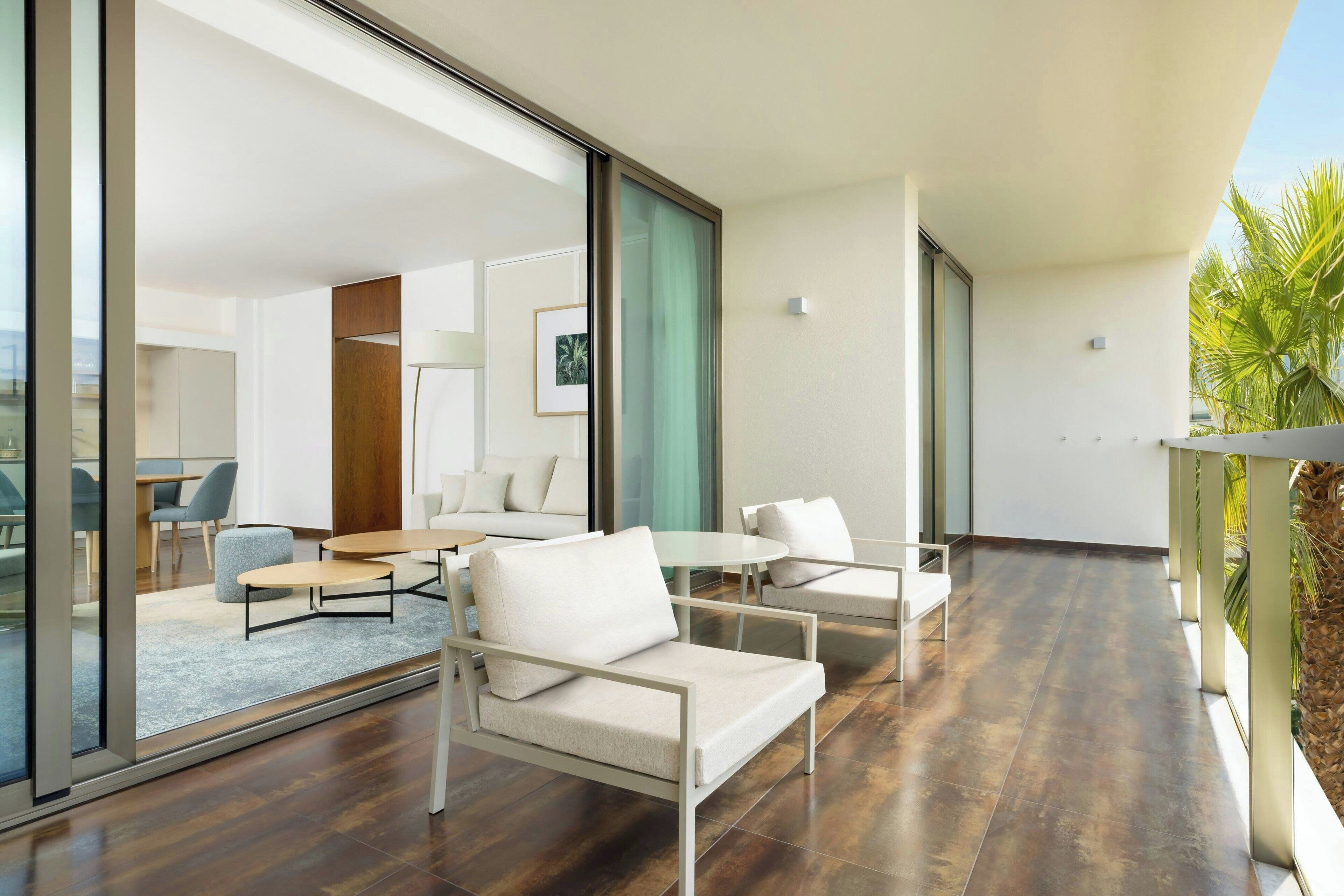 Un intérieur moderne avec un canapé, des fauteuils et une petite table. Il y a une grande fenêtre et une porte qui donne sur une autre pièce.