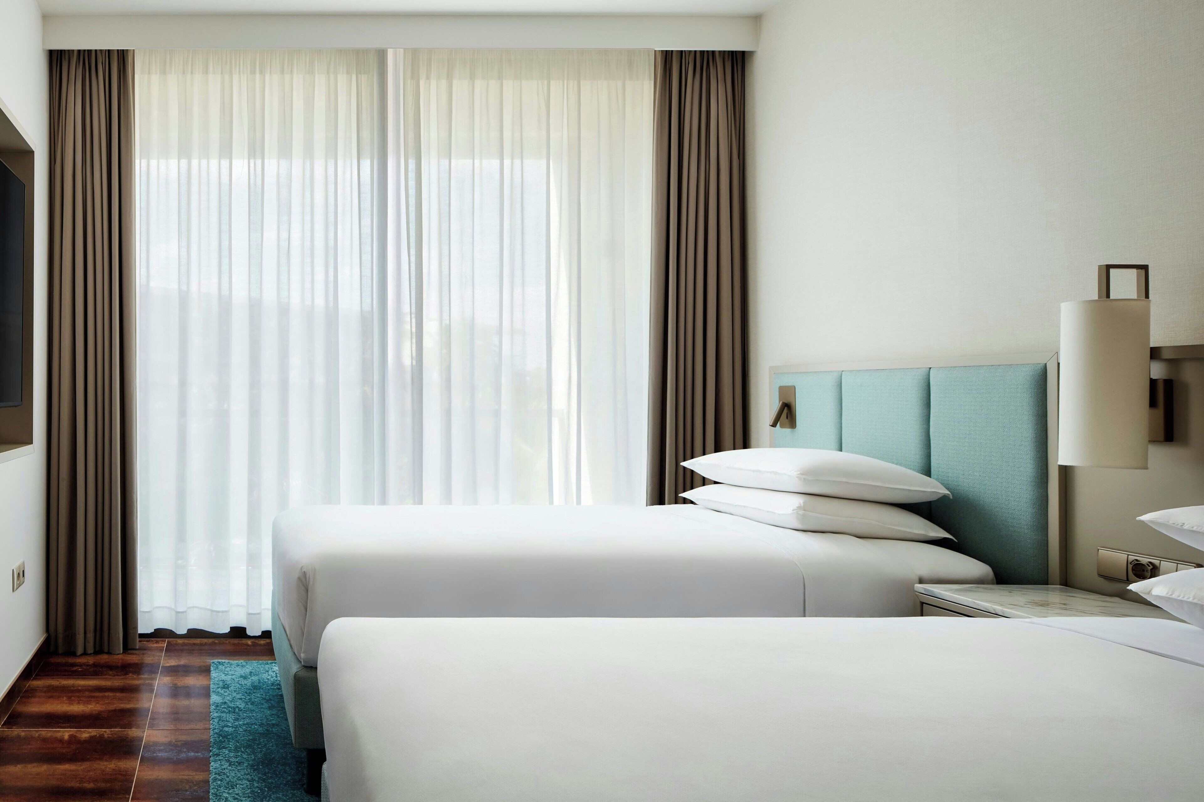 A hotel room with two beds, a large window with sheer curtains, a teal headboard, and a bedside table with a lamp.