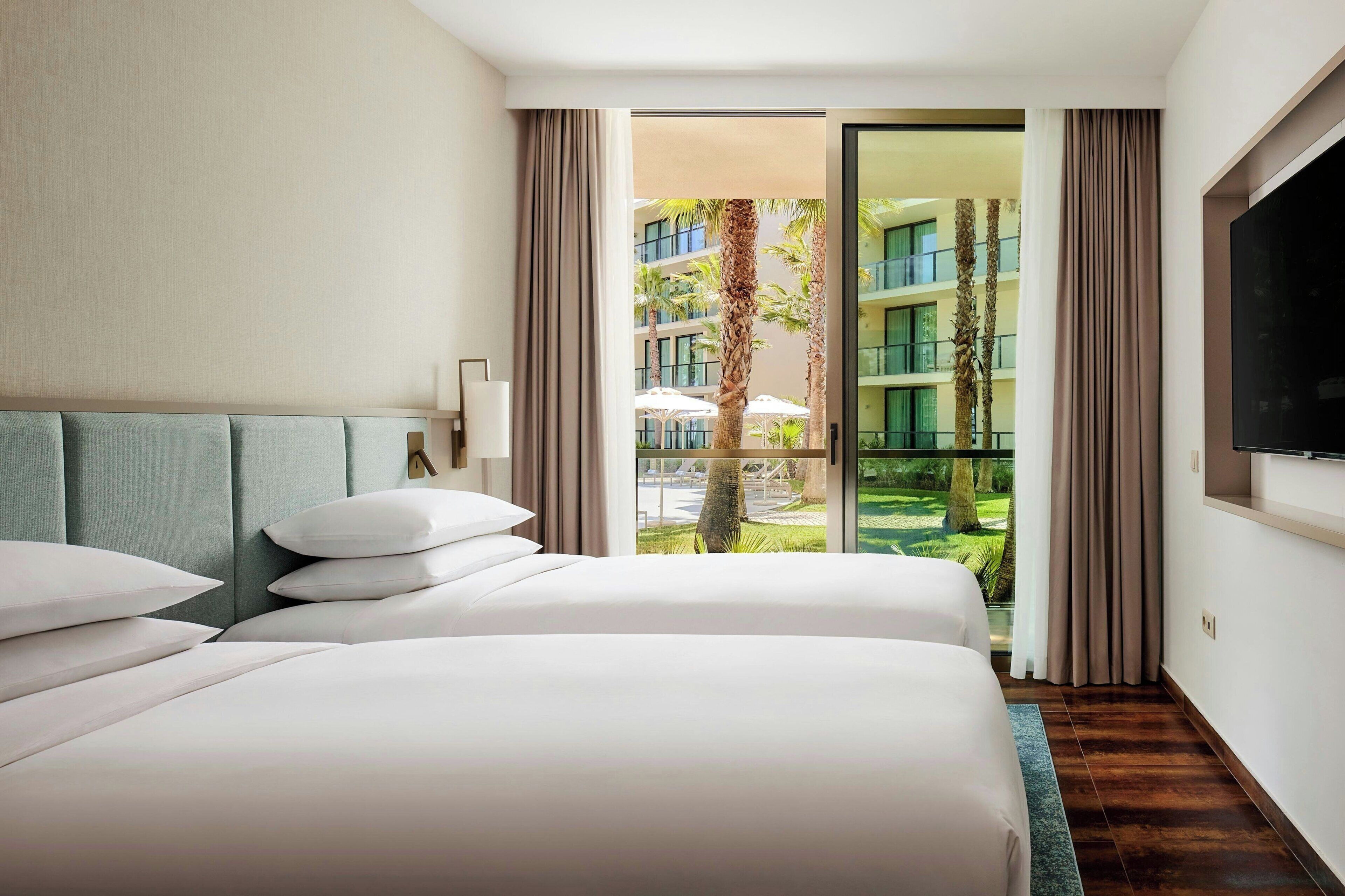 A hotel room with two beds, a large window, and a view of a building and palm trees.