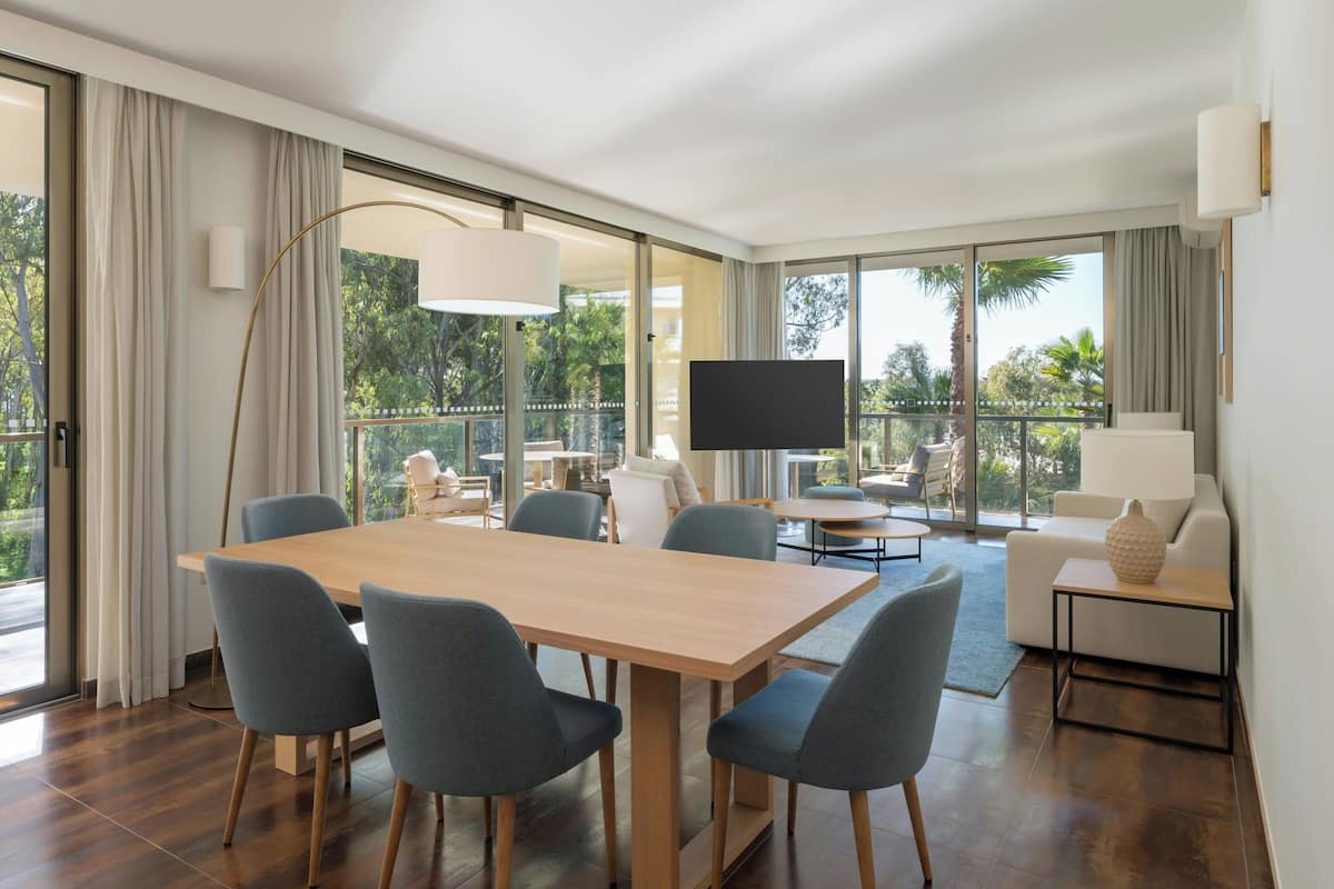 Ein modernes Hotelzimmer mit einem Holztisch, grauen Stühlen, einem Flachbildfernseher, einem Sofa und einem Balkon mit Blick auf Palmen.